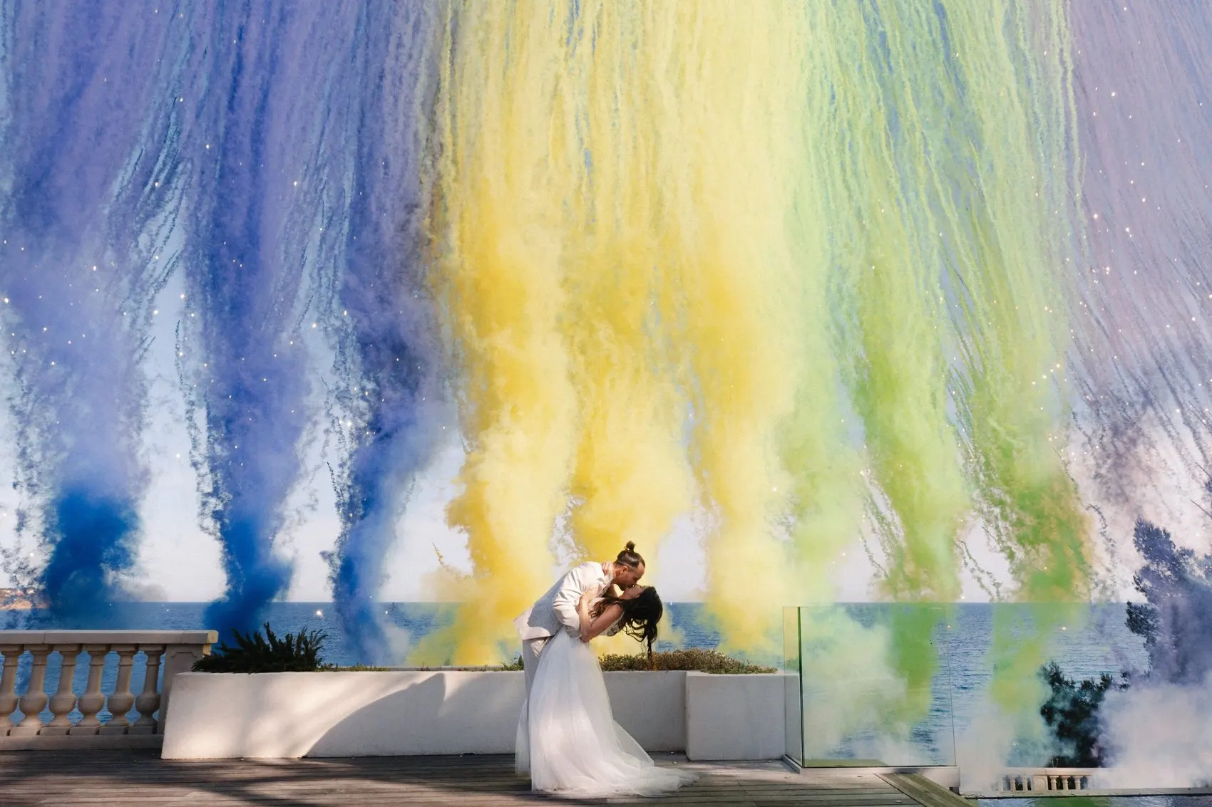 Un couple de mariés s’embrasse devant la mer, sous des fumées colorées.